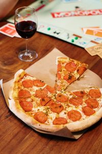 A delicious pepperoni pizza with a glass of red wine beside a board game on a wooden table.