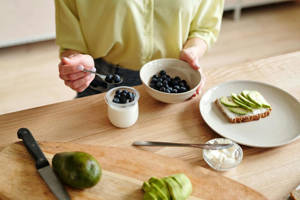 A person prepares a healthy breakfast with yogurt, blueberries, and avocado toast.