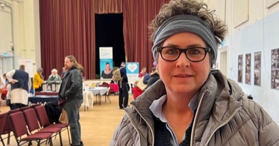 Zannah, a woman wearing a grey coat and grey headband stands in a hall where an event is taking place