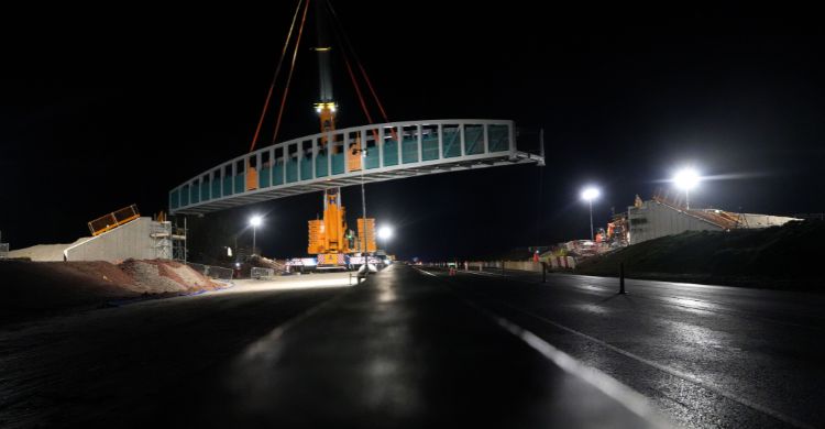 Image shows a steel bridge being lifted by a crane into place above a road at night