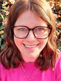 Close up photo of Lauren smiling at the camera. She has shoulder length brown hair and glasses. She is wearing a pink jumper
