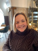Jane is sat in a restaurant type setting. She is smiling at the camera wearing a black jumper and she has a blonde bob hairstyle