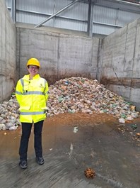Victoria is wearing a hi-viz jacket and hard hat. She is standing in an industrial building with a pile of waste behind her