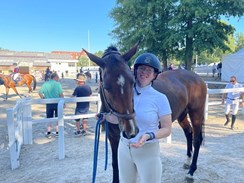 Sarah is standing next to a brown horse who is eating from her hand. She is wearing a white shirt and a black riding hat.
