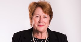Linda is looking at the camera. She is wearing a smart jacket and dress and is wearing a white necklace. She has brown short hair