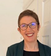Kate is smiling broadly at the camera. She is wearing glasses and has short brown hair, She is wearing a smart jacket and top.