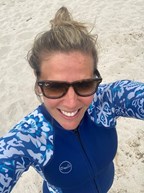 Abigail taking a selfie of herself wearing sunglasses on a beach. She is wearing a blue wetsuit and is smiling