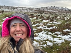 Veryan is taking a selfie with some snow covered rocky ground behind her. Her hood of her coat is up and she is wearing a grey hat underneath. She is smiling.