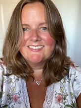 Hollie is smiling at the camera. She has long brown wavy hair and is wearing a white top with flowers on it. She is wearing a silver necklace
