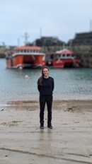 Kimberley is standing on a beach with a harbour and two boats in the background. She is wearing navy blue trousers and jumper.