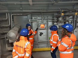 Four women are wearing hi-viz jackets and hard hats in an industrial setting. Three of the women are looking at one woman who is standing near a steel door with a window showing a furnace inside