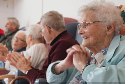 A woman in her nineties is pictured smiling and holding her hands with 4 other older people in the background talking to each other